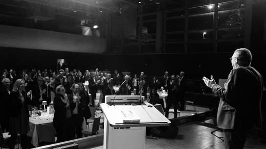Pressemitteilung: Verabschiedung Dr. Tobias Böcker Standing Ovation bei der Verabschiedung von Dr. Tobias Böcker, als er auf der Bühne steht und ins Publikum blickt.
